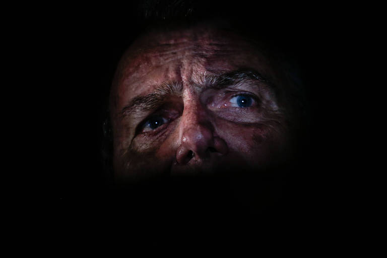 A imagem mostra um close-up do rosto de Jair Bolsonaro, com foco em seus olhos azuis e expressões faciais marcadas. O fundo é predominantemente escuro, destacando apenas a parte superior do rosto e os olhos, que transmitem uma sensação de intensidade e mistério.
