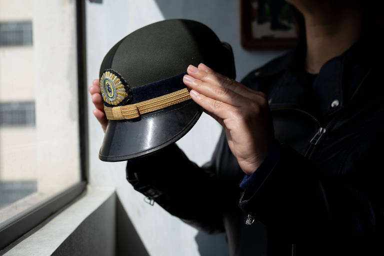 Mulher branca vestida com farda segura cap preto, verde-oliva e dourado que compõe o uniforme