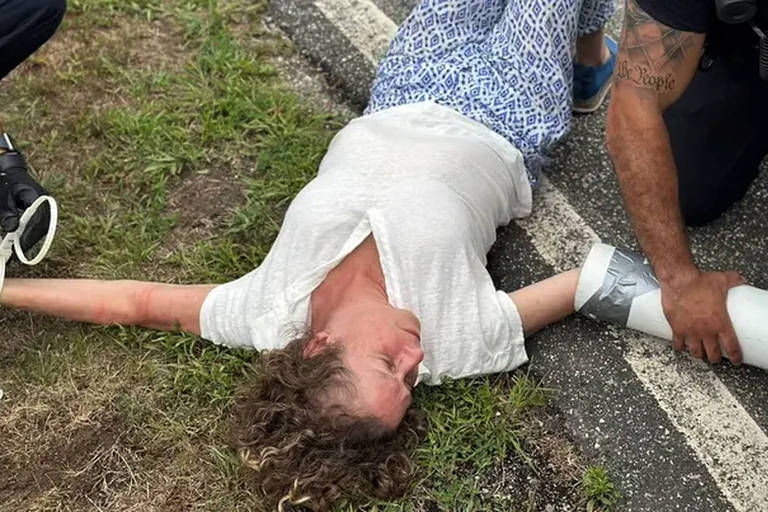 Mulher deitada na margem de pista de asfalto, com parte do corpo sobre a grama, e braços abertos. É possível ver braços de agentes de segurança que tentam desatar cordas e outros materiais que prendem os punhos dela aos de outros ativistas; Abigail é branca e tem cabelos claros 