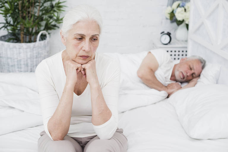 Homem idoso dormindo e mulher idosa sentada na cama com a expressão de desânimo