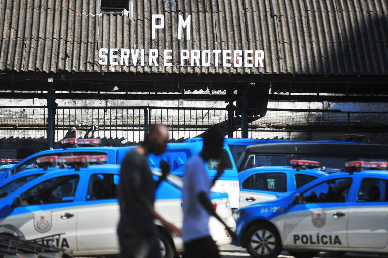 Pessoas passam em frente a batalhão da Polícia Militar que diz em letreiro: PM Servir e proteger