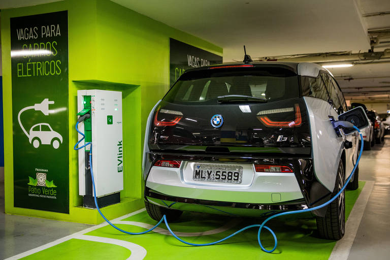 Imagem mostra carro elétrico sendo carregado por meio de um fio conectado a uma fonte. Ele está em um estacionamento, numa área pintada de verde.