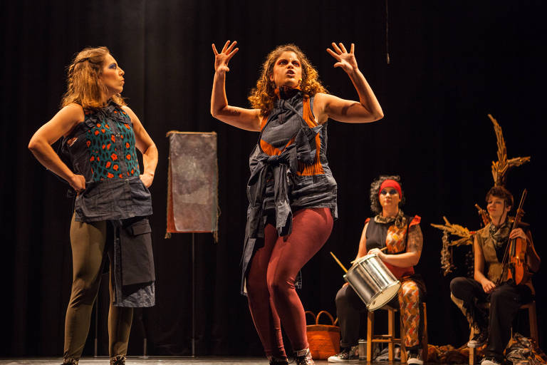 A imagem mostra duas mulheres em destaque no palco, uma à esquerda com cabelo liso e castanho claro, vestindo uma blusa com estampa de onça e calças verdes, e a outra à direita com cabelo cacheado e castanho, usando uma blusa azul escura e calças vermelhas, gesticulando expressivamente. Ao fundo, há três mulheres sentadas: uma com cabelo encaracolado e faixa vermelha, outra com cabelo curto e uma blusa colorida, e a terceira tocando um violino. O cenário é escuro, com um pano de fundo preto e um objeto decorativo ao centro.