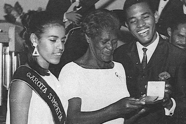 Foto em preto e branco traz ao centro Laudelina de Campos Mello, uma senhora preta, que usa vestido longo claro, recebendo uma condecoração do cantorJair Rodrigues, à sua esquerda. Ele é  um homem negro, alto e sorridente. Usa terno e gravata. À direita de Laudelina, uma modelo preta, também de vestido branco, usa uma faixa diagonal sobre o corpo. 