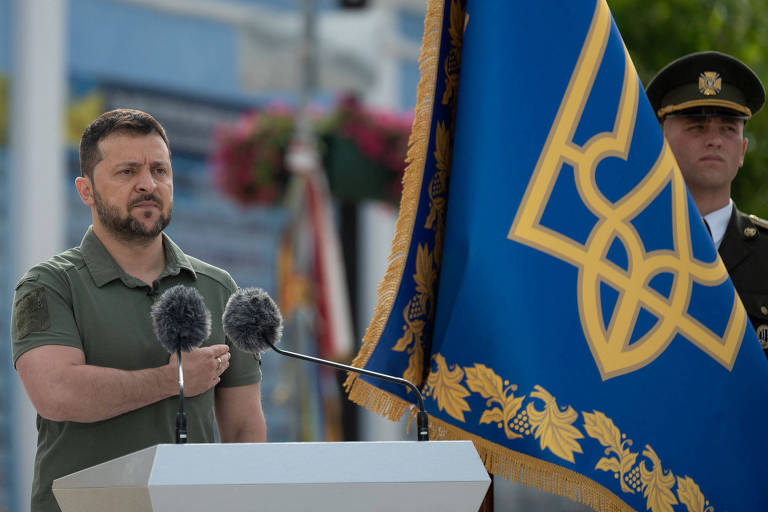 Homem de camisa pólo verde-militar coloca mão sob o peito; ele está atrás de um púlpito com microfones e de uma bandeira ucraniana