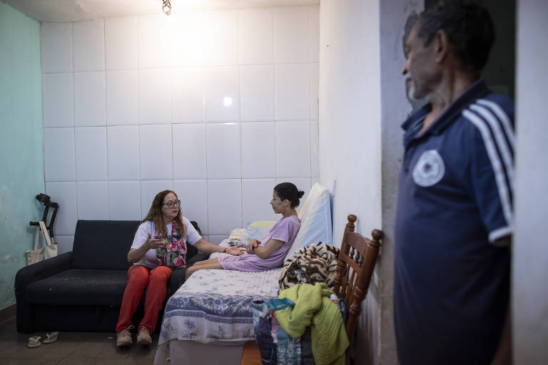 Moradora voluntária conversa com paciente em cuidados paliativos atendida por comunidade compassiva na Rocinha, no Rio de Janeiro . Ambas estão sentadas e são observadas por um homem em pé.