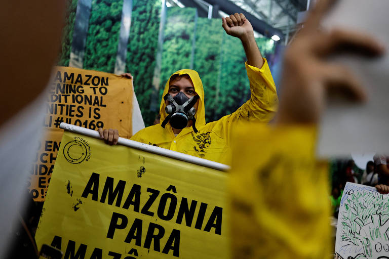 Entre cartazes, homem usa máscara contra gases tóxicos e capa de chuva amarela; ele segura cartaz onde é possível ler Amazônia para Amazônia