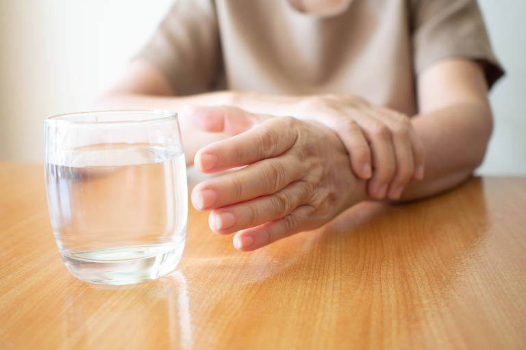 Mãos de mulher idosa com sintoma de tremor alcançando um copo de água na mesa de madeira. As causas do tremor nas mãos incluem doença de Parkinson, acidente vascular cerebral ou lesão cerebral. Distúrbio neurológico de saúde mental.