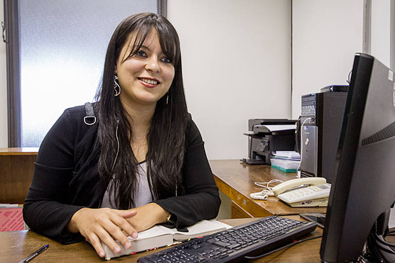 Imagem colorida mostra uma mulher branca, de cabelos longos, sentada e mexendo em um teclado de computador