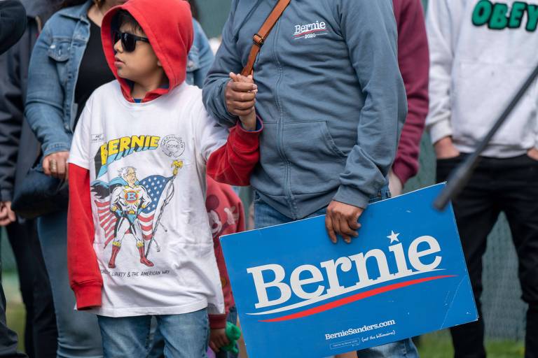 Garoto branco com óculos escuros, casaco de capuz e, por cima, camiseta branca com desenho de super herói e a inscrição "super bernie"; ao lado, adulto branco de blusa azul com pasta com a inscrição Bernie