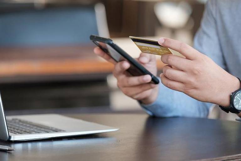 Pessoa com cartão de crédito e celular na mão em frente a um laptop
