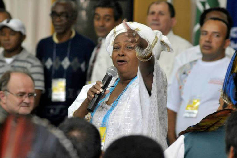 Mulher negra com roupa branca segura microfone com uma mão e aponta para a frente com a outra; ela está cercada por outras pessoas