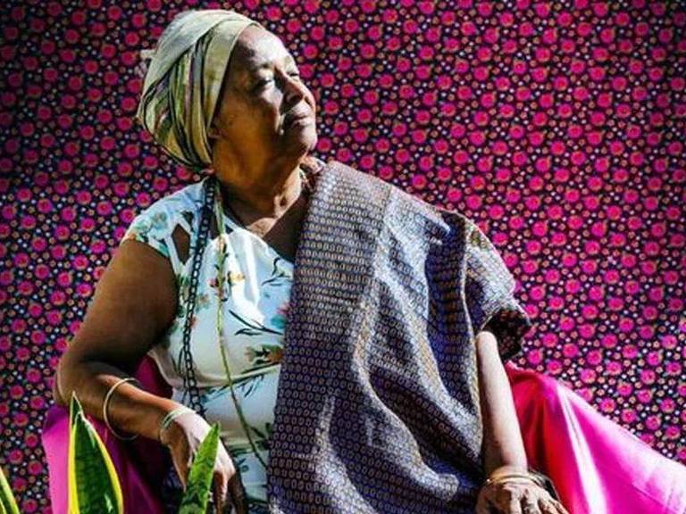 Foto mostra mulher negra sentada em uma cadeira, Ela usa um turbante e veste uma roupa colorida