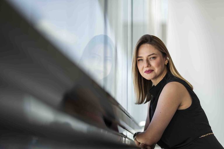 A presidente da SulAmérica, Raquel Reis, aparece na foto apoiada na janela do escritório. Não é possível ver a paisagem pela janela. Só os vidros. Ela está de lado, vestida de preto. Tem cabelos loiros 