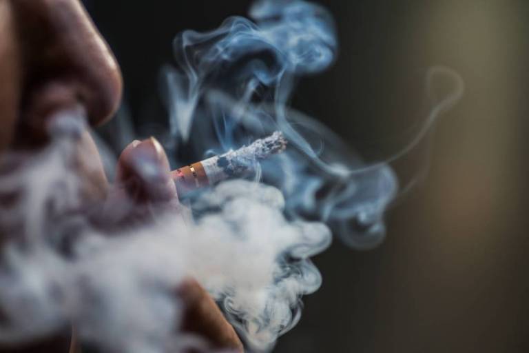Fotografia foca a mão de uma pessoa segurando um cigarro e soltando a fumaça pela boca