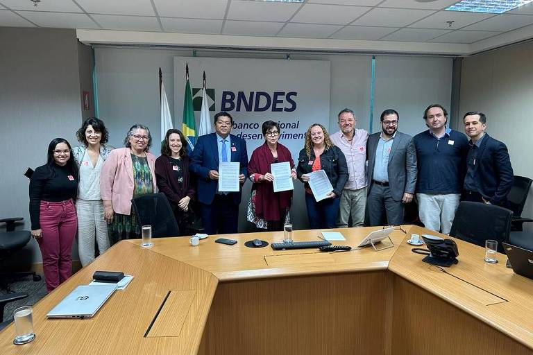 Em uma sala de escritório, com mesa em forma do de letra U, 11 pessoas aparecem em pé frente à sala e posam para a foto sorrindo. Entre homens e mulheresm todos vestem roupas formais como blazers, caças e, no caso das mulheres, saias e também calças. Três deles seguram nas mãos um documento. Ao fundo há um telão para exibição de slides onde se lê "BNDES"