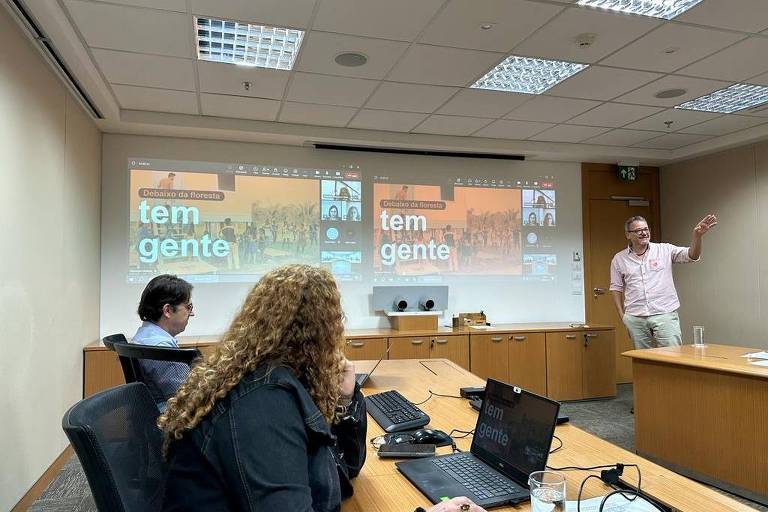 Em uma sala de reunião, um homem realiza uma apresentação de slides a um grupo de pessoas. Na foto, outras duas pessoas assistem à apresentação