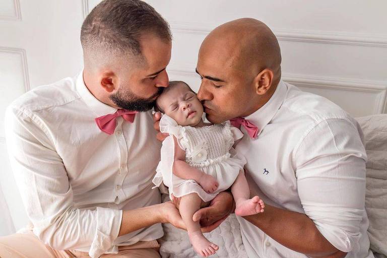 dois homens de branco e gravata borboleta beijam bebê recem-nascida ao centro