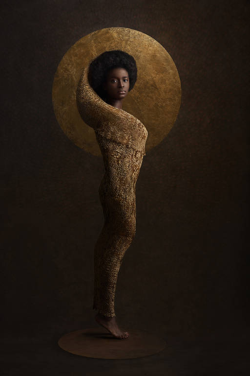 A imagem retrata uma mulher negra, ela usa um cabelo afro e um traje feito de folhas douradas