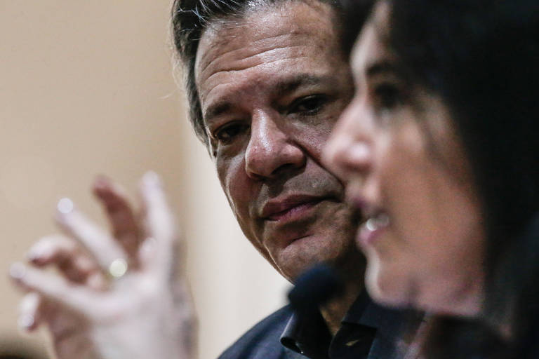 O ministro Fernando Haddad tem os olhos baixos e a testa franzida. Usa roupas pretas. Tebet, em primeiro plano, está desfocada, e gesticula com as mão enquanto fala.