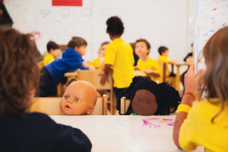 Na imagem há duas bonecas, uma preta e uma branca; elas estão sentadas em cadeiras de uma sala de aula, ao fundo aparecem alunos também sentados