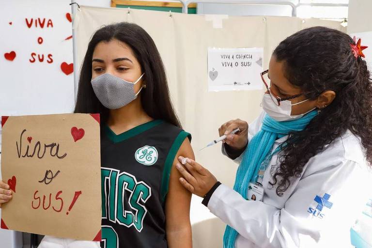 Uma adolescente é vacinada por uma profissional de saúde do SUS. A jovem segura uma placa com a mensagem "Viva o SUS"