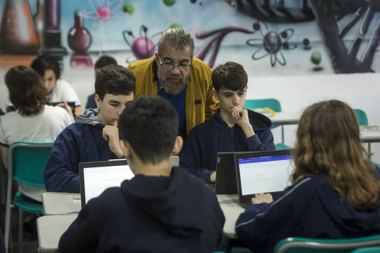 O professor Marcelo Cavalcante está em pé, orientando alunos sentados à mesa com computadores em uma sala de aula