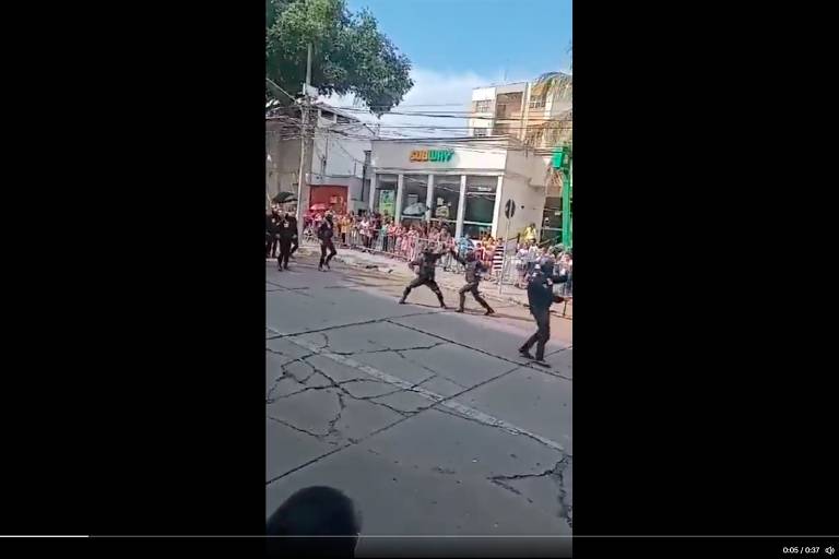 Pessoas vestidas de uniforme escuro parecem lutar, com os braços se encontrando no alto. Estão a céu aberto, desfilando, com pessoas assistindo à cena atrás de grade. Ao fundo, uma unidade do restaurante Subway e céu azul