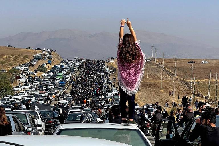 Mulher sem véu em cima de veículo em estrada no Irã diante de pessoas a caminho de cemitério após morte de Mahsa Amini, em 2022