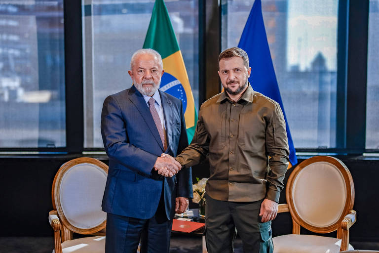Lula está à esquerda e, Zelenski, à direita. Os dois estão em pé, se cumprimentando, em ambiente fechado. Há bandeiras de seus respectivos países atrás deles e janelas