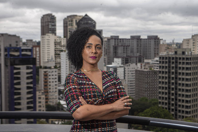 Mulher negra posa para foto séria e com os braços cruzados. Ao fundo se vê prédios.