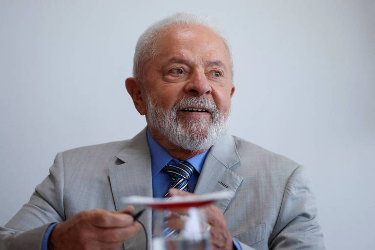 Lula durante reunião no Palácio do Planalto. Ele veste terno cinza, camisa e gravata azul. Lula sorri e olha para o lado direito da imagem. Ele aparece do peito para cima