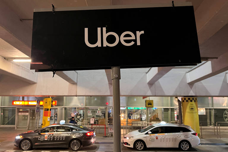 Imagem mostra placa de publicidade preta com logo da Uber na cor branca. É possível ver dois carros estacionados no  espaço, um preto e outro branco, em frente a uma entrada do aeroporto de Varsóvia, na Polônia. 