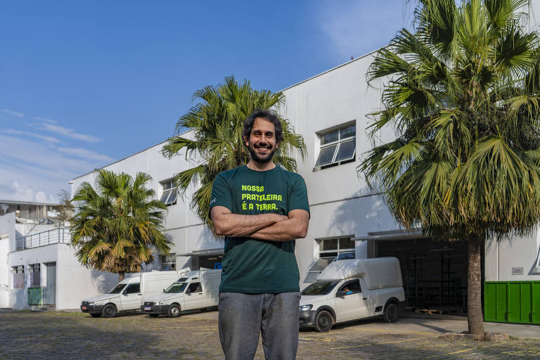 Tomás é um homem branco de cabelos castanhos, ele usa uma camisa na qual está escrito 'nossa prateleira é a terra'; ele está de pé, em frente à sede da empresa, ao fundo está o prédio branco e três carros de transporte