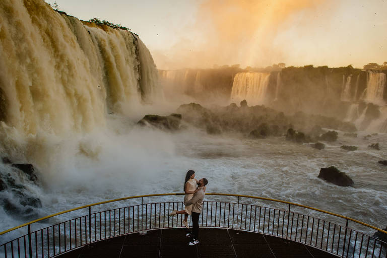 homem pega mulher no colo em mirante com cataratas ao fundo e nascer do sol