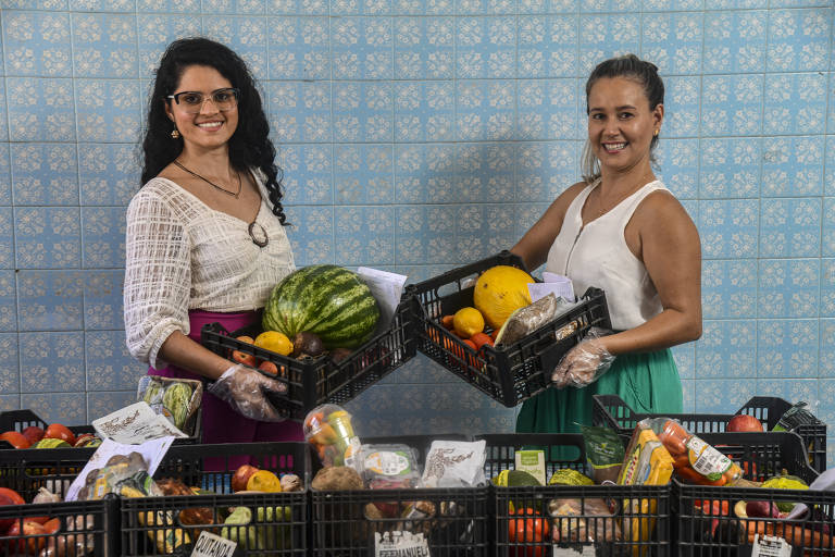 Hany e Manuela seguram cestas com produtos orgânicos, como melancia, cenoura, tomate, batata, maça, laranja, abacate e melão; Hany é uma mulher branca de cabelos loiros presos em um rabo de cavalo, ela veste camisa branca e calça verde; Manuela é uma mulher branca, de cabelos pretos e cacheados, ela veste uma camisa branca e uma calça rosa