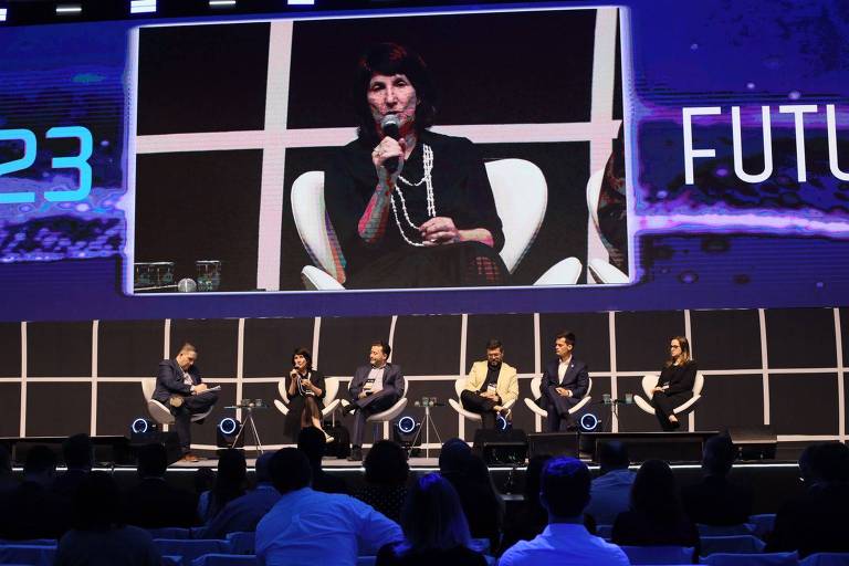 Seis debatedores sentados num palco, embaixo de um grande telão focado em uma das expositoras