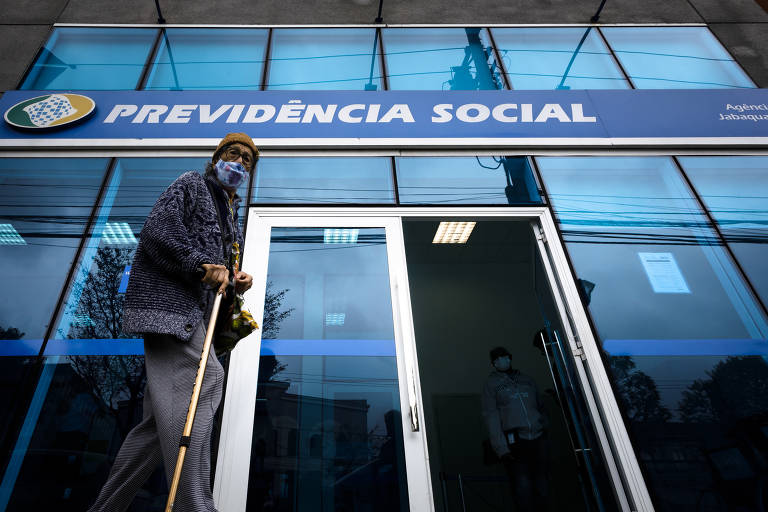 Senhora idosa usando máscara e gorro amarelo, andando com auxílio de uma bengala na mão direita na frente da fachada da Previdência Social