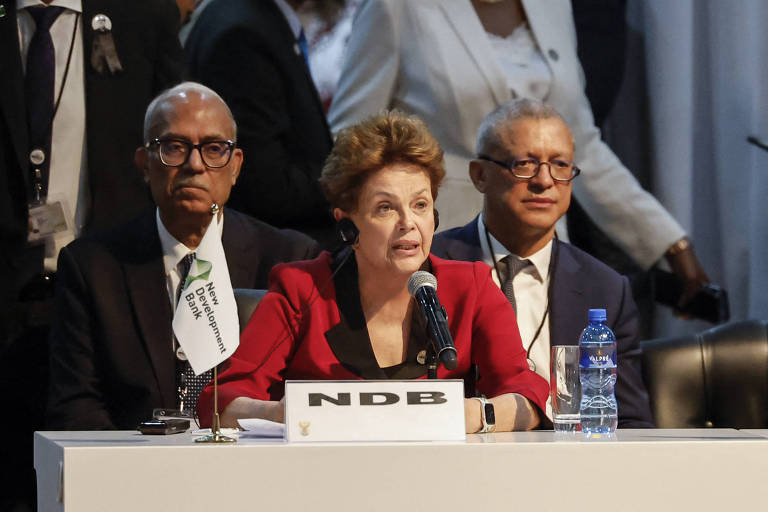 Dilma, mulher branca, de camisa preta e blazer vermelho, fala ao microfone, sentada em uma mesa com placa branca com a sigla NBD escrita em preto. Ao fundo, dois homens sentados