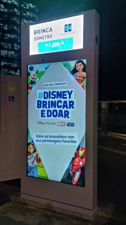 Um painel luminosos na vertical. No alto: Brinquedômetro e o valor de R$1200,00. No centro, dentro de um losângo #Disney Brincar e doar, entre na brincadeira com seus personagens favoritos.  Nas extremidades do losango os personagens Mickey, Moana, Mirabel e Homem de Ferro