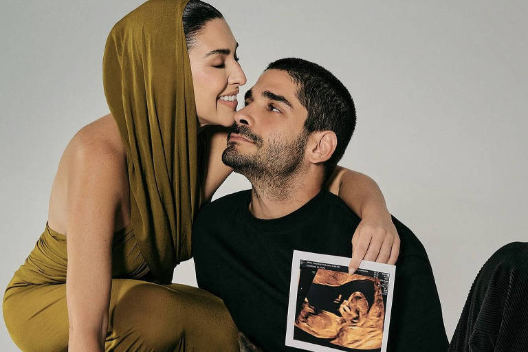 Uma mulher branca usando um vestido de cor mostarda está agachada, abraçando um homem também branco, de roupas pretas. Ela segura na mão uma imagem de um ultrassom que mostra um bebê. Junto a eles, está um cachorro grande deitado no chão.