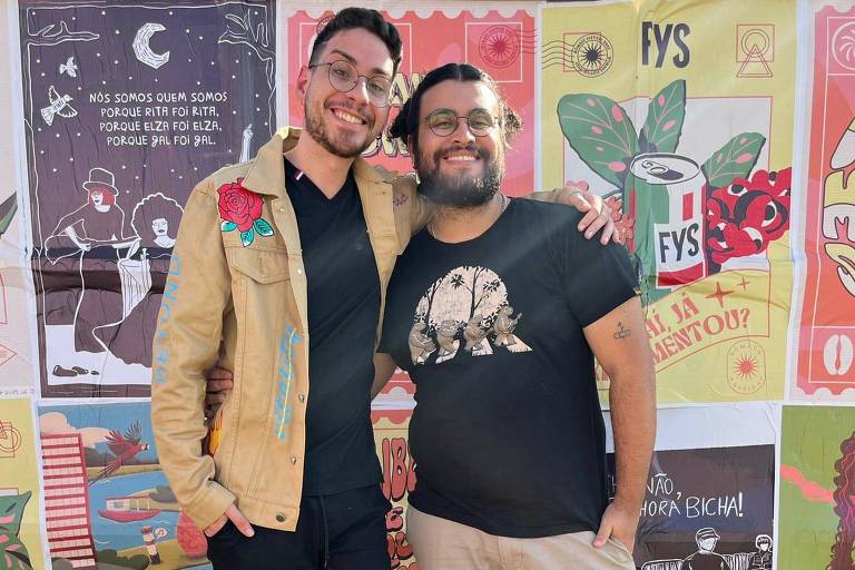 Fotografia colorida mostra o casal Eric Munir e Vinicius Maranhão; eles são brancos de cabelos escuros, estão de pé, abraçados, e sorrindo para a câmera