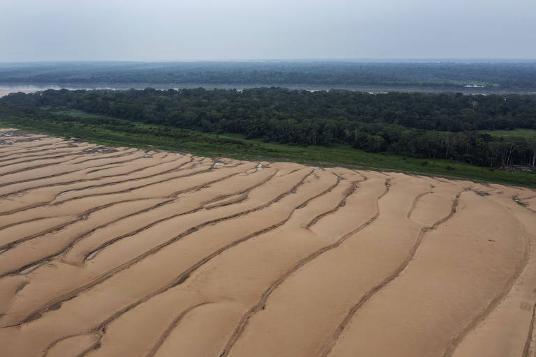 A imagem mostra uma vista aérea do leito seco do rio, com várias linhas paralelas de solo arenoso exposto. Ao fundo, há uma faixa de vegetação densa e uma linha de água.
