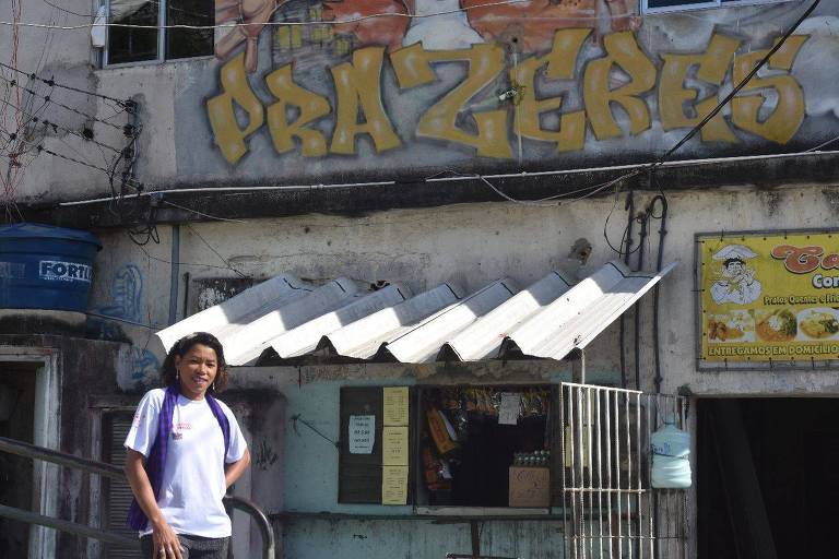 Em área externa de favela, mulher negra posa para a foto frente a um muro com grafite que diz "Prazeres" em referência à favela carioca