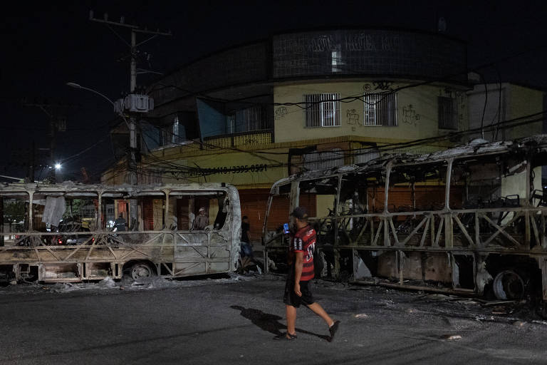 homem com camisa do flamengo passa duas carcaças de onibus incendiada em rua à noite