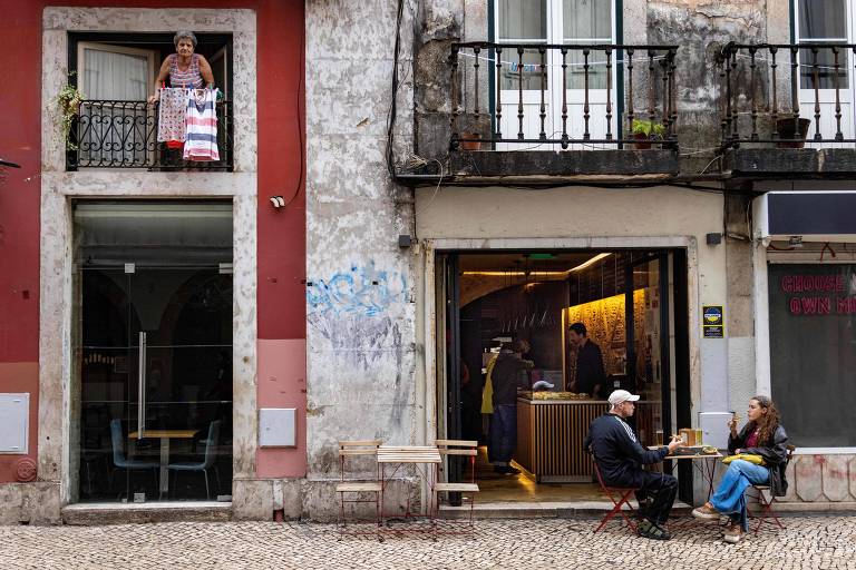 Uma mulher pendura a roupa enquanto as pessoas saboreiam uma refeição na calçada em frente a uma cafeteria na Rua de São Paulo, em Lisboa, Portugal