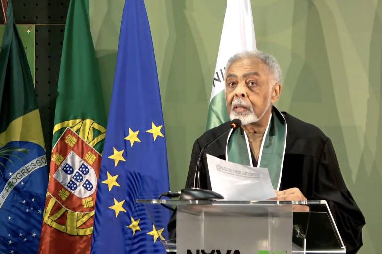 O cantor e compositor Gilberto Gil recebeu título de doutor honoris causa pela Universidade Nova de Lisboa. Ele, que é negro, aparece na foto no pódio, enquanto discursa. Ele traja a capa preta dos doutores e tem uma fita verde no pescoço. Gil está em frente às bandeiras do Brasil, de Portugal e da União Europeia