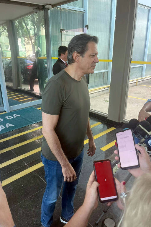 Haddad de calça jeans e camiseta básica verde oliva fala em pé em saguão de prédio. Mãos seguram celulares