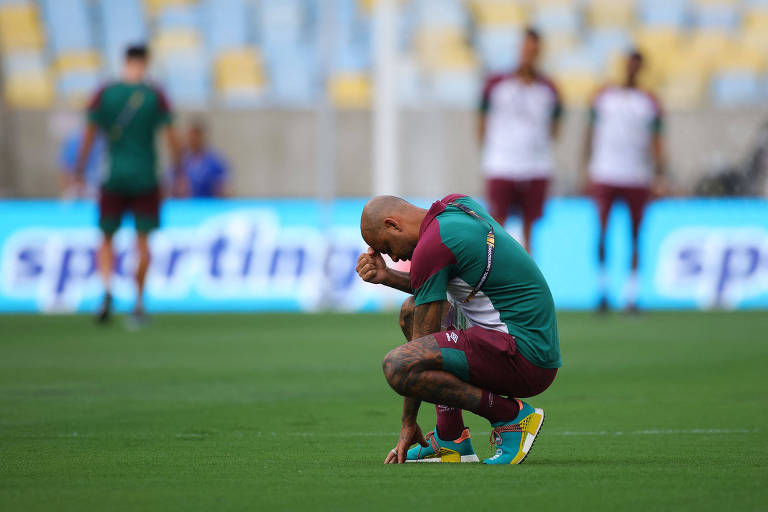 Melo, um homem negro e careca, está agachado no gramado. Uma mão dele toca a grama e a outra segura sua cabeça.