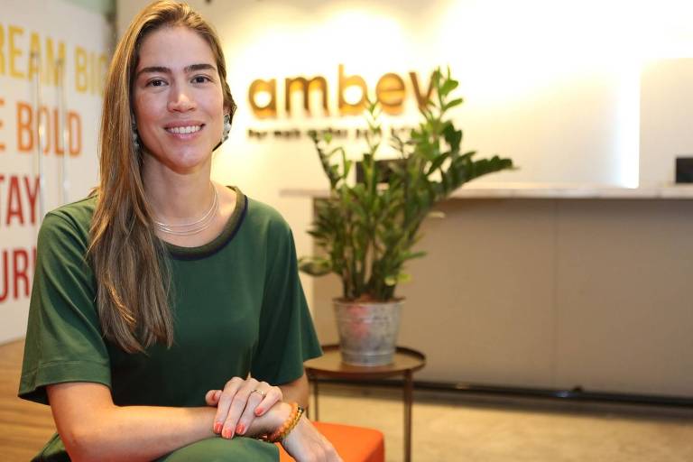 Foto mostra mulher sentada e sorrindo para a câmera. Local é escritório corporativo com a logo marca da Ambev em uma das paredes ao fundo. A mulher é branca, loira e magra. usa uma camista de cor verde escuro e calça de mesma cor